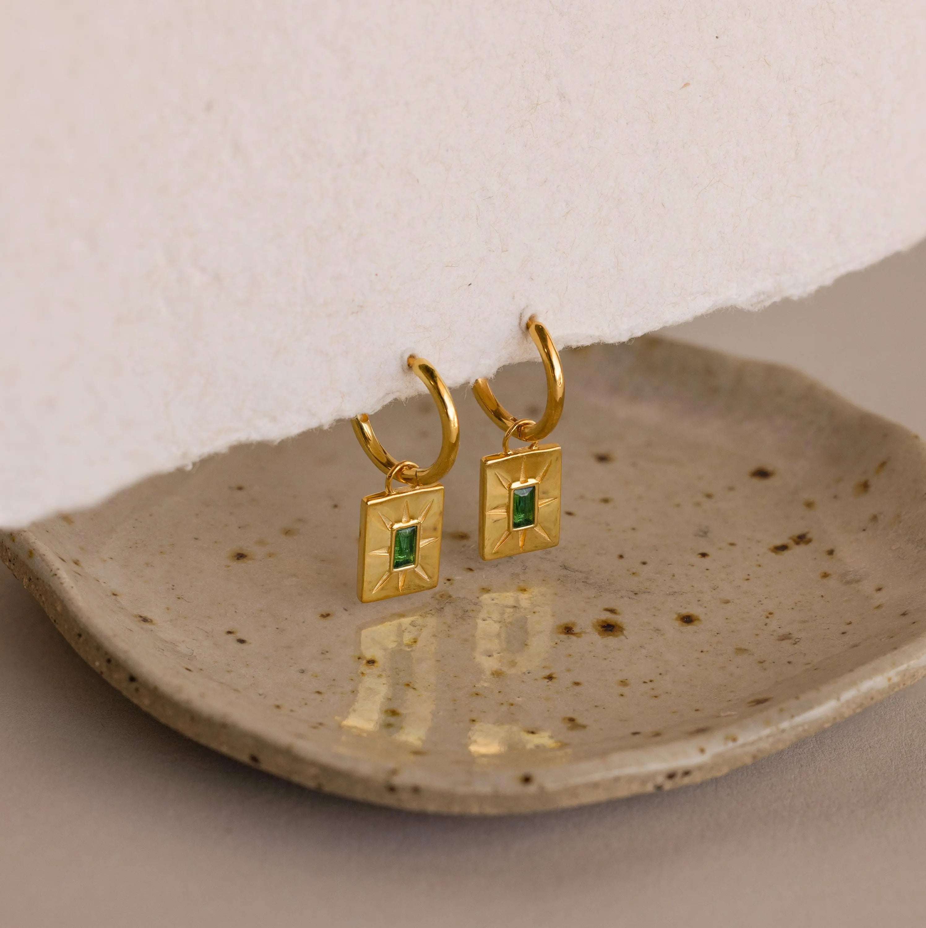 Birthstone Tag Huggies, gold hoops with rectangular green stones, elegantly displayed on a textured ceramic dish.
