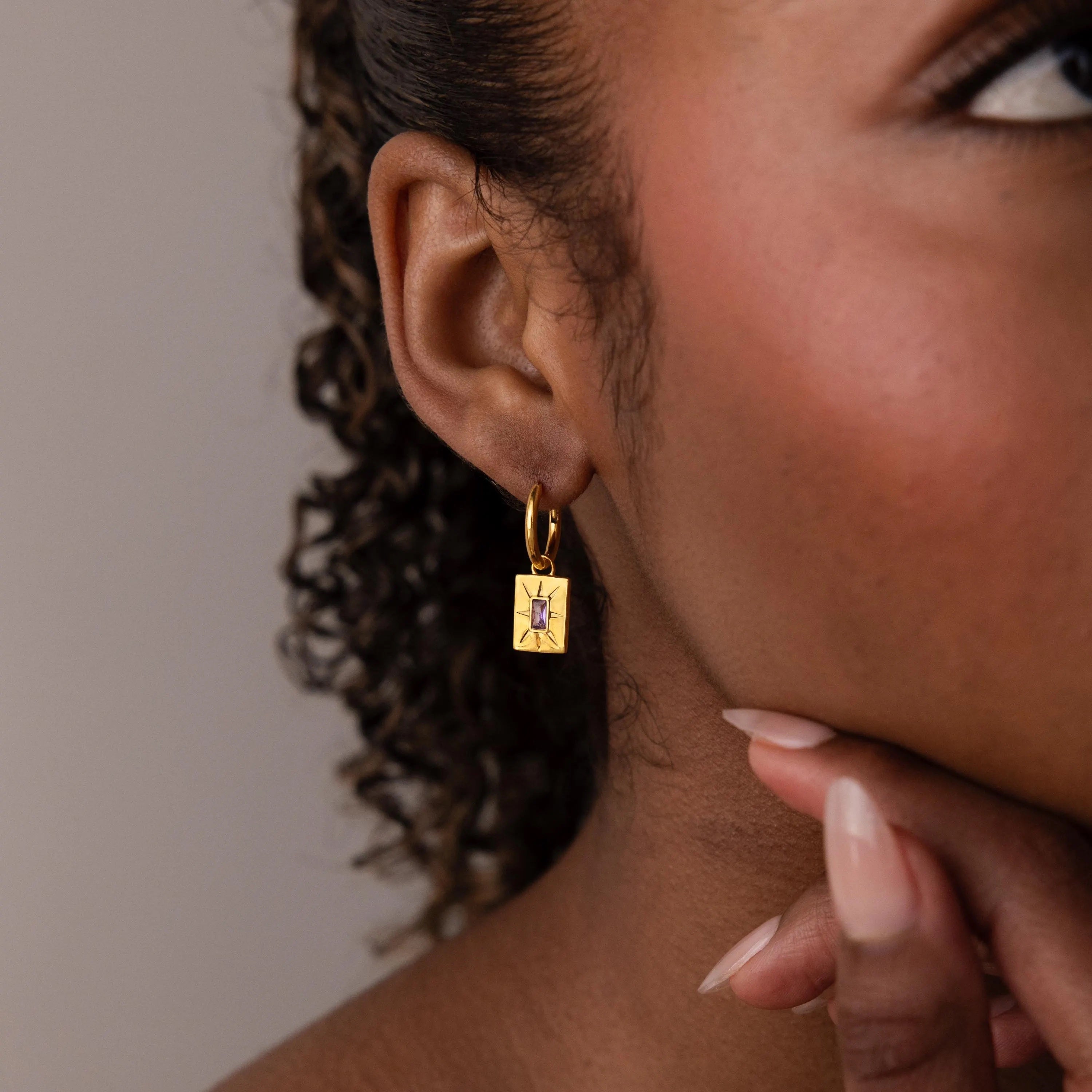 Close-up of a woman wearing Birthstone Tag Huggies, gold rectangular hoops featuring a purple star design, with her hand resting on her chin.