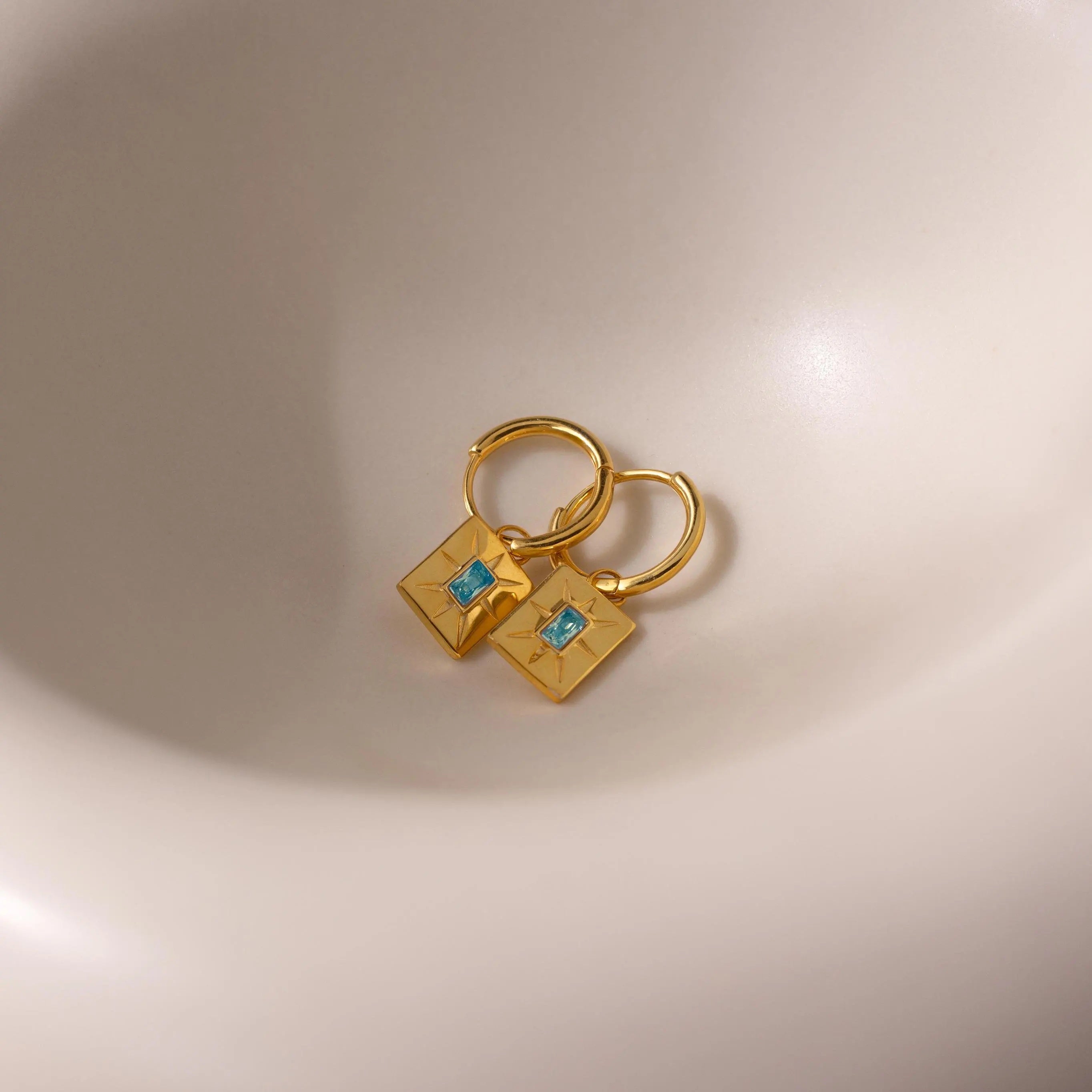 Birthstone Tag Huggies: Gold huggie hoops with square pendants and small blue stones, displayed on a smooth light surface—ideal personalized jewelry for adding a birthstone-inspired touch to your style.