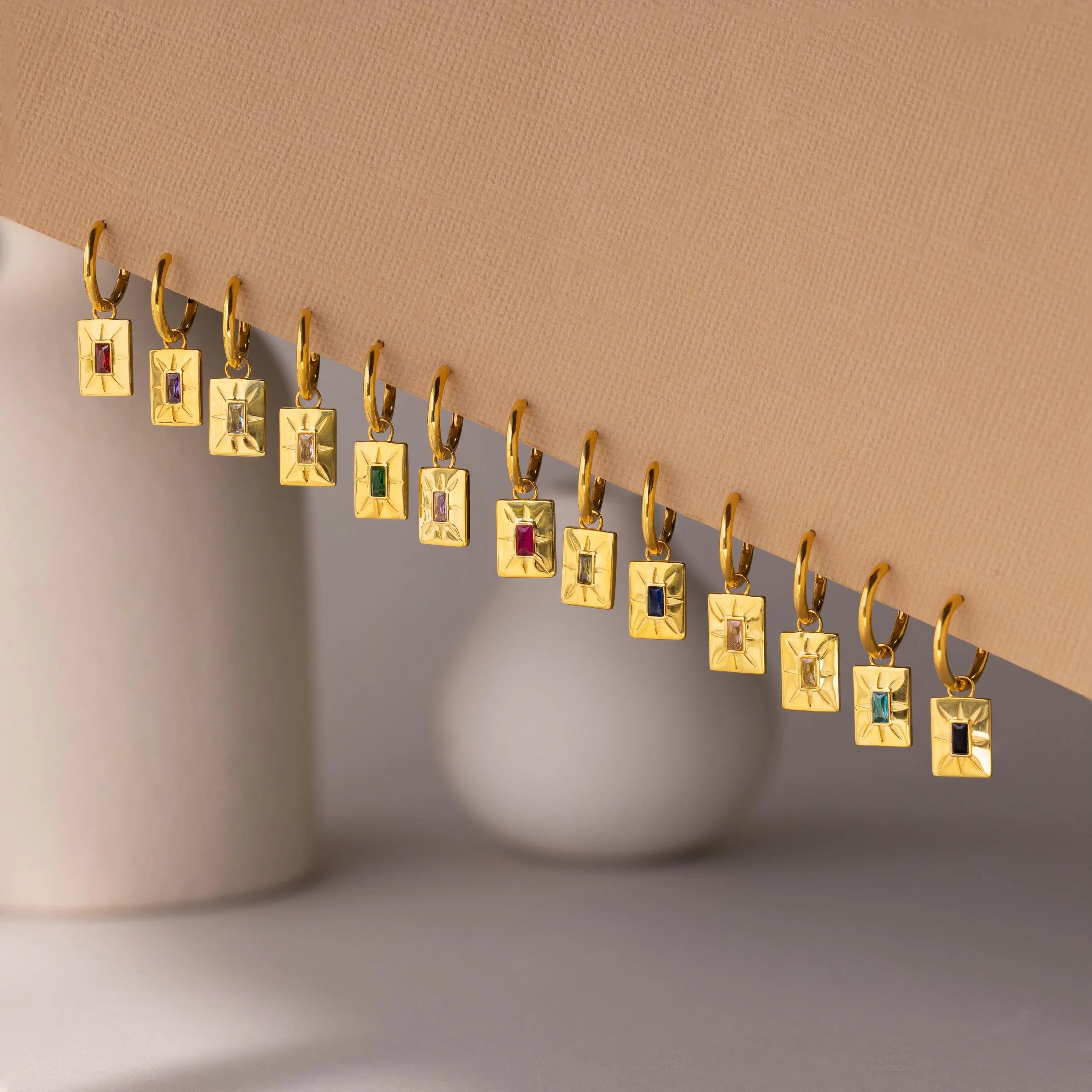 Birthstone Tag Huggies—gold rectangular pendants with vibrant stones—are shown in a row on a slanted surface against a neutral background, ideal for pairing with other huggie hoops or personalized jewelry.