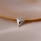 The Shark Tooth Charm in Sterling Silver sits on a smooth, light surface beside a beige shell—a sea-inspired jewelry accent.