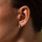 Close-up of a woman's ear wearing a small, smooth green jade stud earring with a gold halo setting.