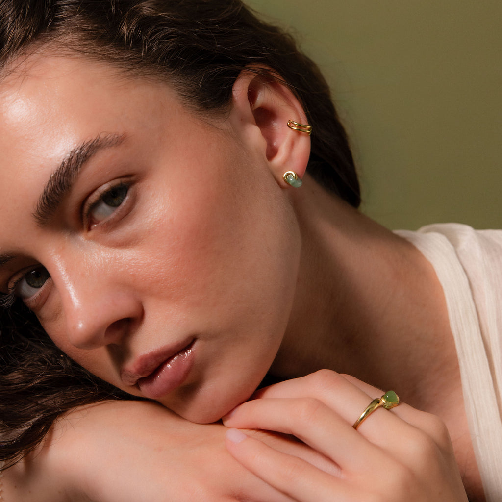 Woman wearing small gold stud earrings with smooth green jade drops, a gold ear cuff, and a matching jade ring.