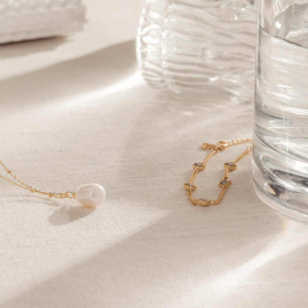 The Venus Diamond Station Bracelet rests on cream fabric beside a glass of water and a ribbed vase—an elegant choice for a wedding look or as an anniversary gift.
