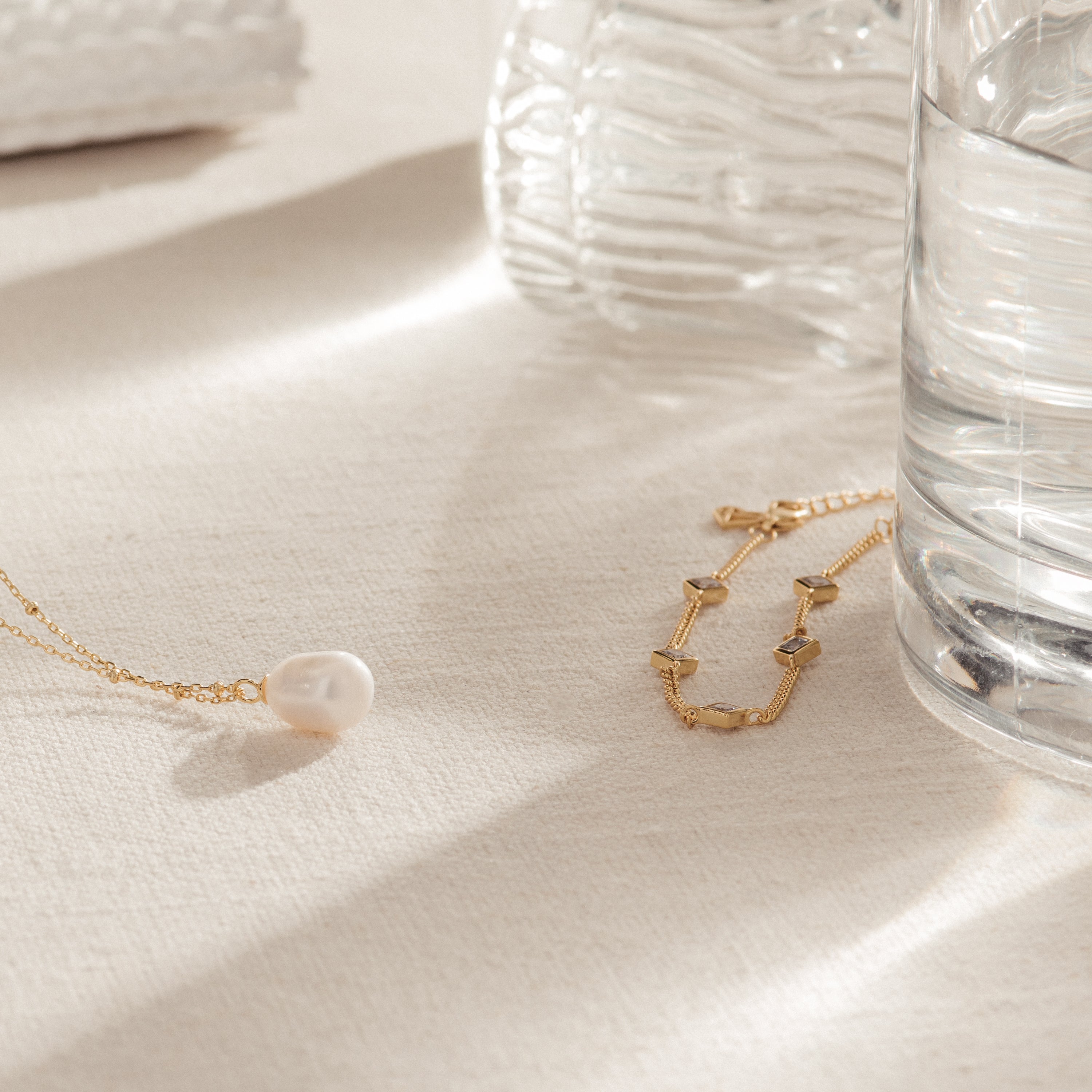 The Venus Diamond Station Bracelet rests on cream fabric beside a glass of water and a ribbed vase—an elegant choice for a wedding look or as an anniversary gift.