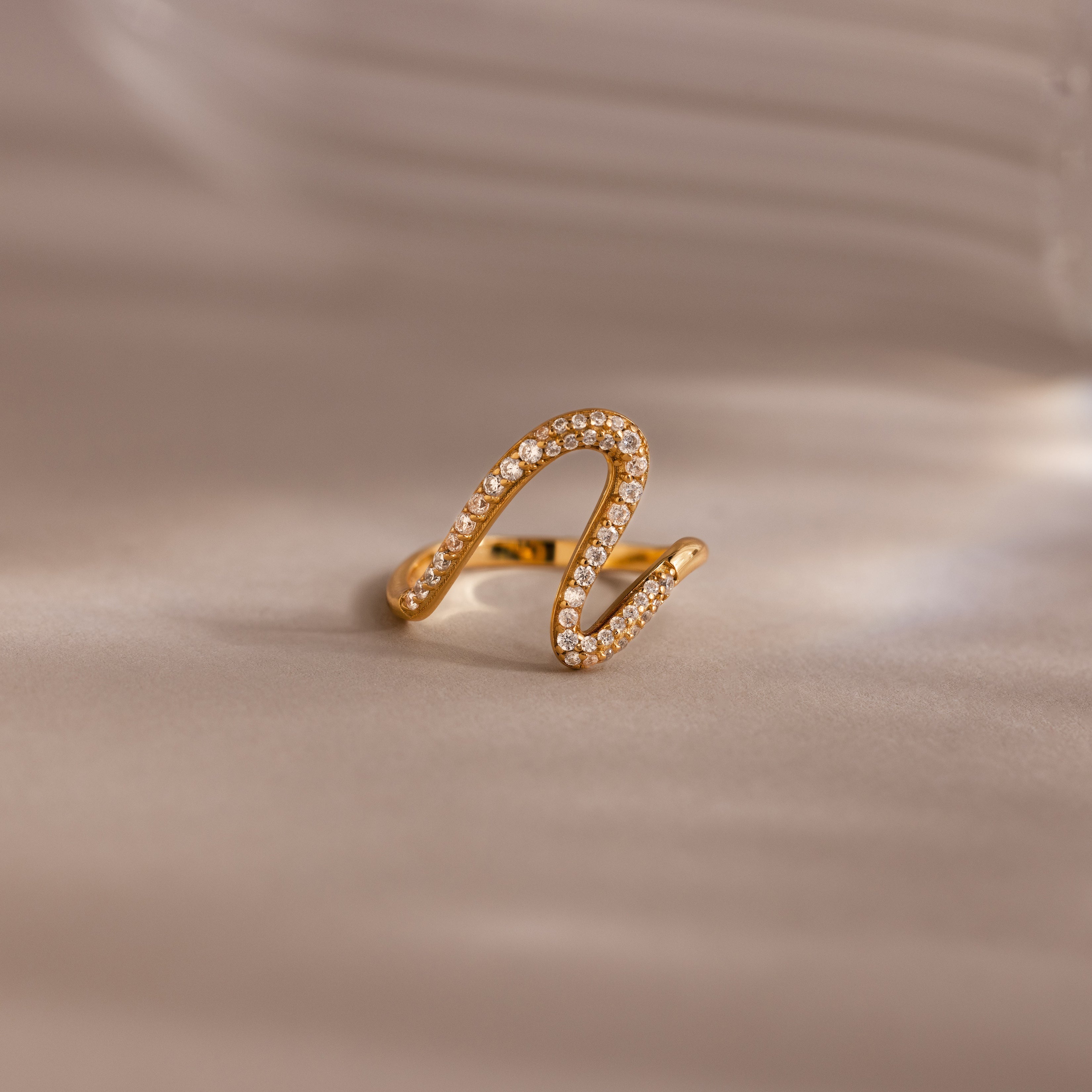 The Pave Wave Ring, featuring a gold wavy design encrusted with small diamonds, is shown on a soft beige background—ideal for stacking and adding subtle elegance to any look.