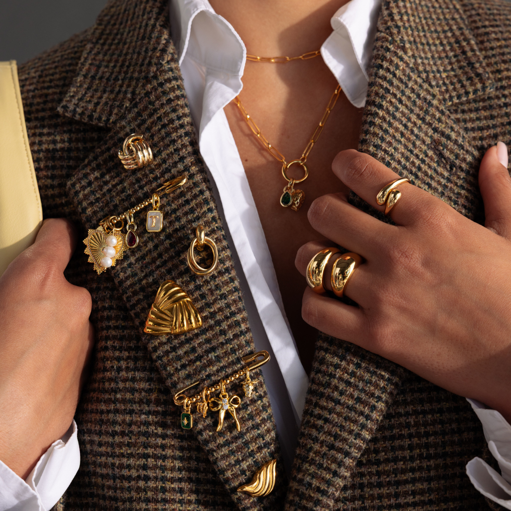 A person wears a brown plaid blazer decorated with multiple gold brooches, including a personalized pin and the Beaded Charm Pin, along with rings and layered necklaces.