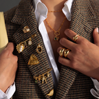 A person wears a brown plaid blazer decorated with multiple gold brooches, including a personalized pin and the Beaded Charm Pin, along with rings and layered necklaces.