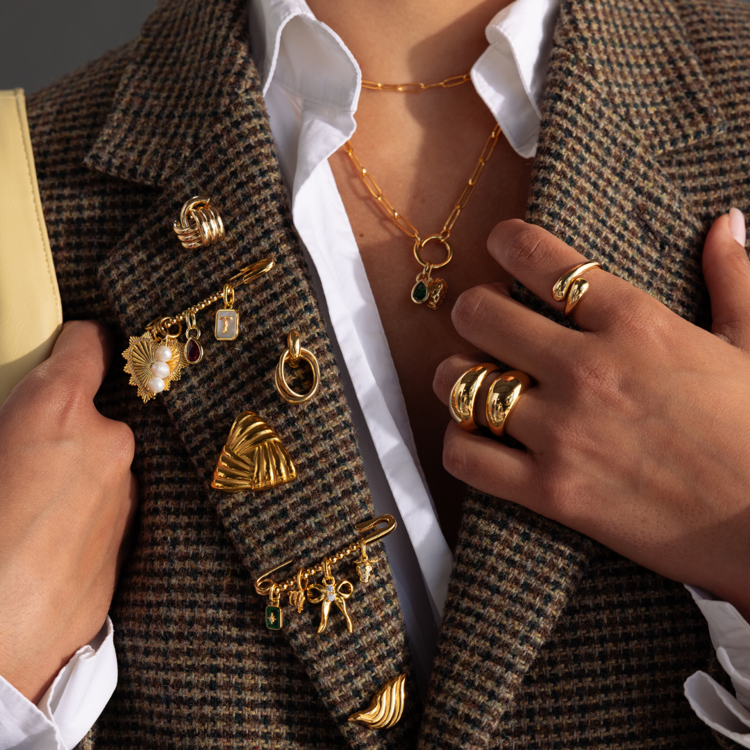 A person wears a brown plaid blazer decorated with multiple gold brooches, including a personalized pin and the Beaded Charm Pin, along with rings and layered necklaces.
