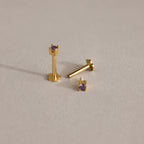 Three Dainty Birthstone Flatback Studs with small purple gemstones are shown on a neutral, textured surface, perfect for multiple piercings.
