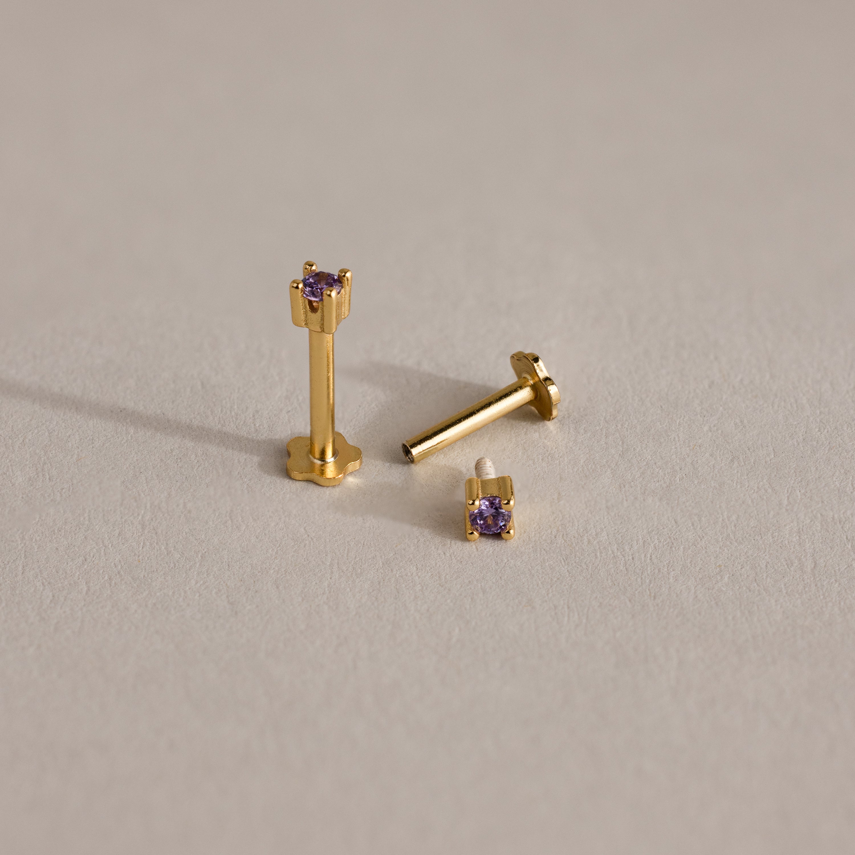 Three Dainty Birthstone Flatback Studs with small purple gemstones are shown on a neutral, textured surface, perfect for multiple piercings.