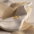 The Dainty Birthstone Flatback Studs are timeless gold earrings with clear gemstones, ideal for multiple piercings, and shown on soft cream-colored fabric.