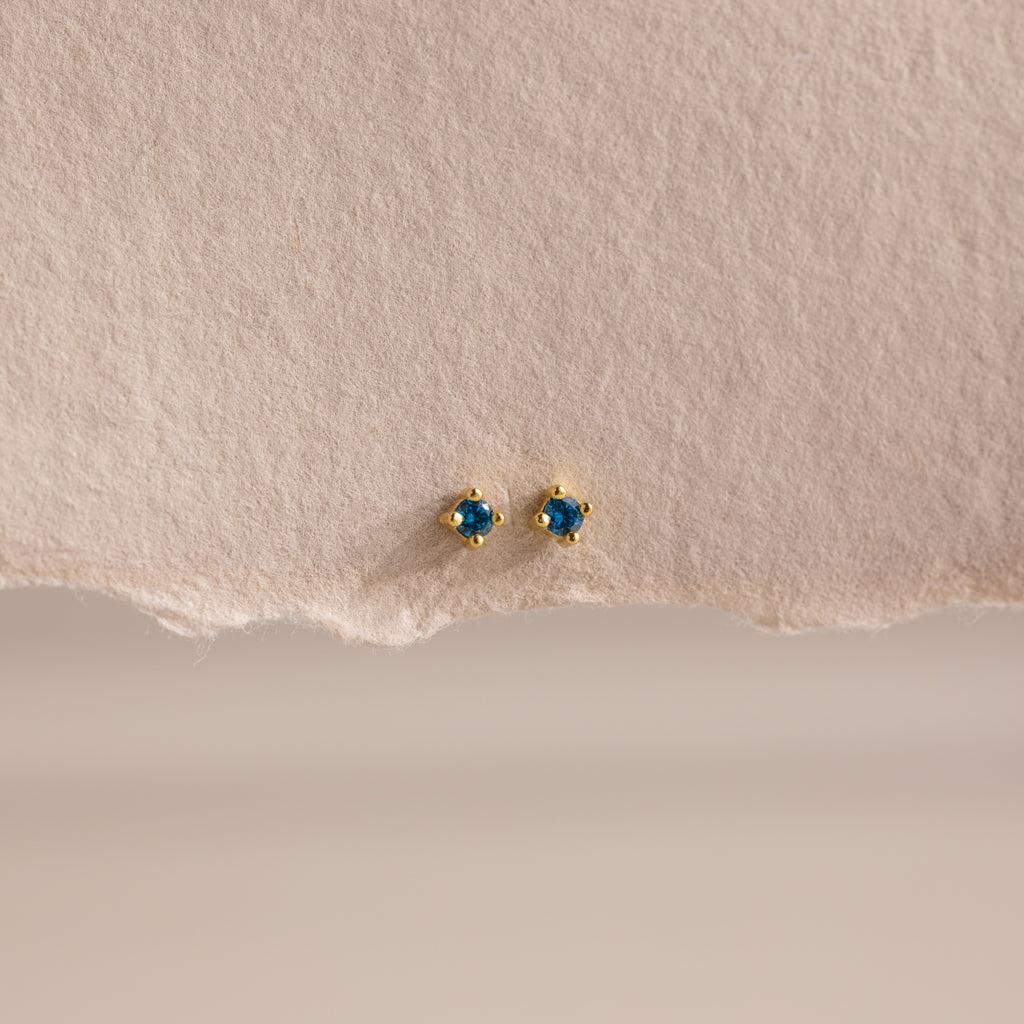 Dainty Blue Zircon Flatback Studs with petite gold settings and vibrant blue zircon stones, ideal for multiple piercings, showcased on a textured beige paper background.