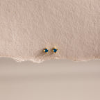 Dainty Blue Zircon Flatback Studs with petite gold settings and vibrant blue zircon stones, ideal for multiple piercings, showcased on a textured beige paper background.