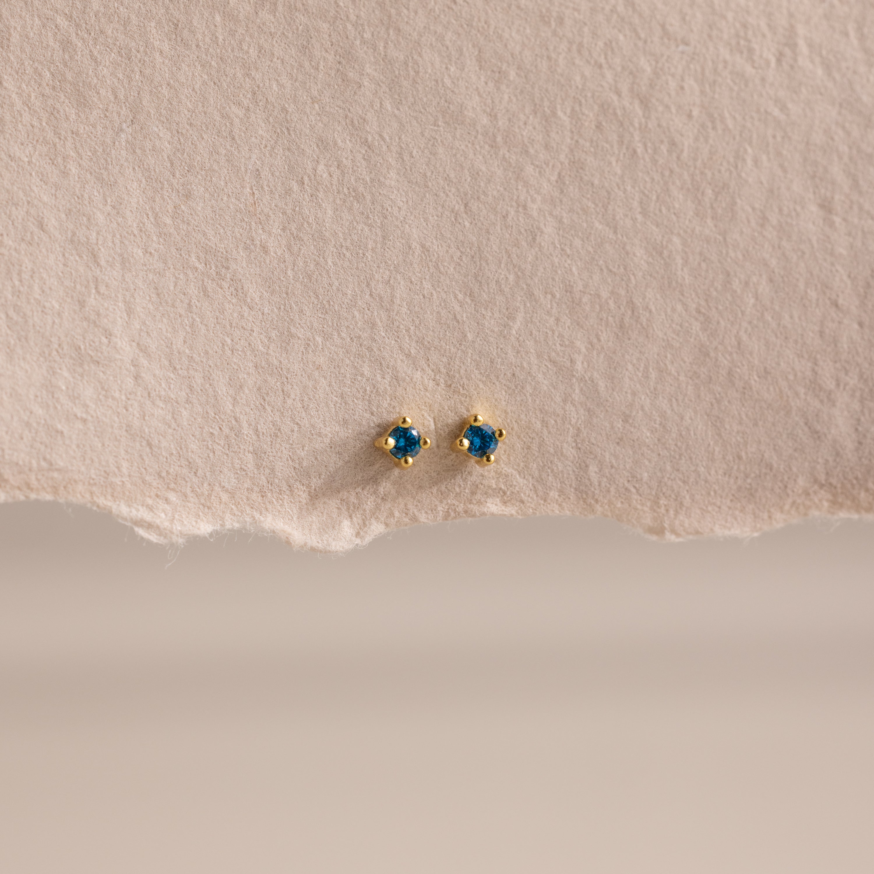 Dainty Blue Zircon Flatback Studs with petite gold settings and vibrant blue zircon stones, ideal for multiple piercings, showcased on a textured beige paper background.