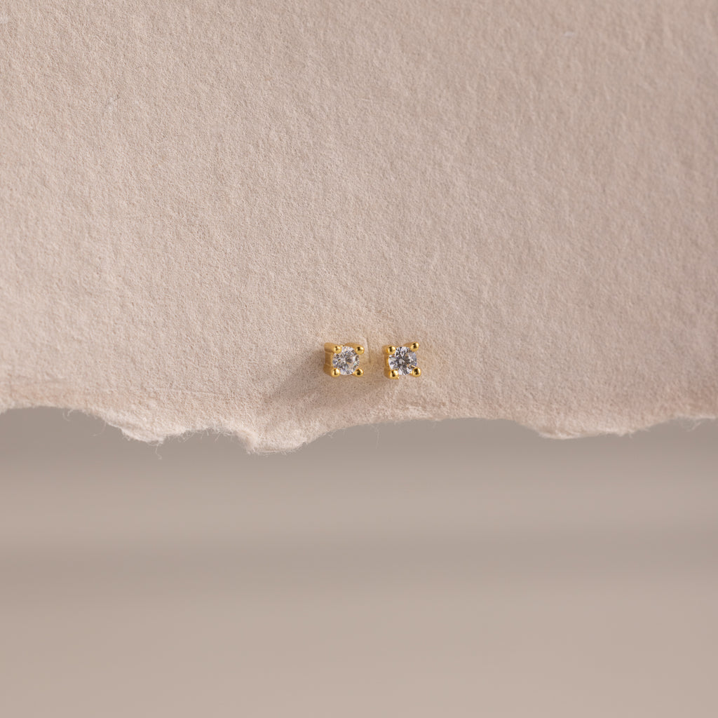 The Dainty Diamond Flatback Studs feature clear gemstones, ideal for multiple piercings, and are shown on a textured beige paper background.