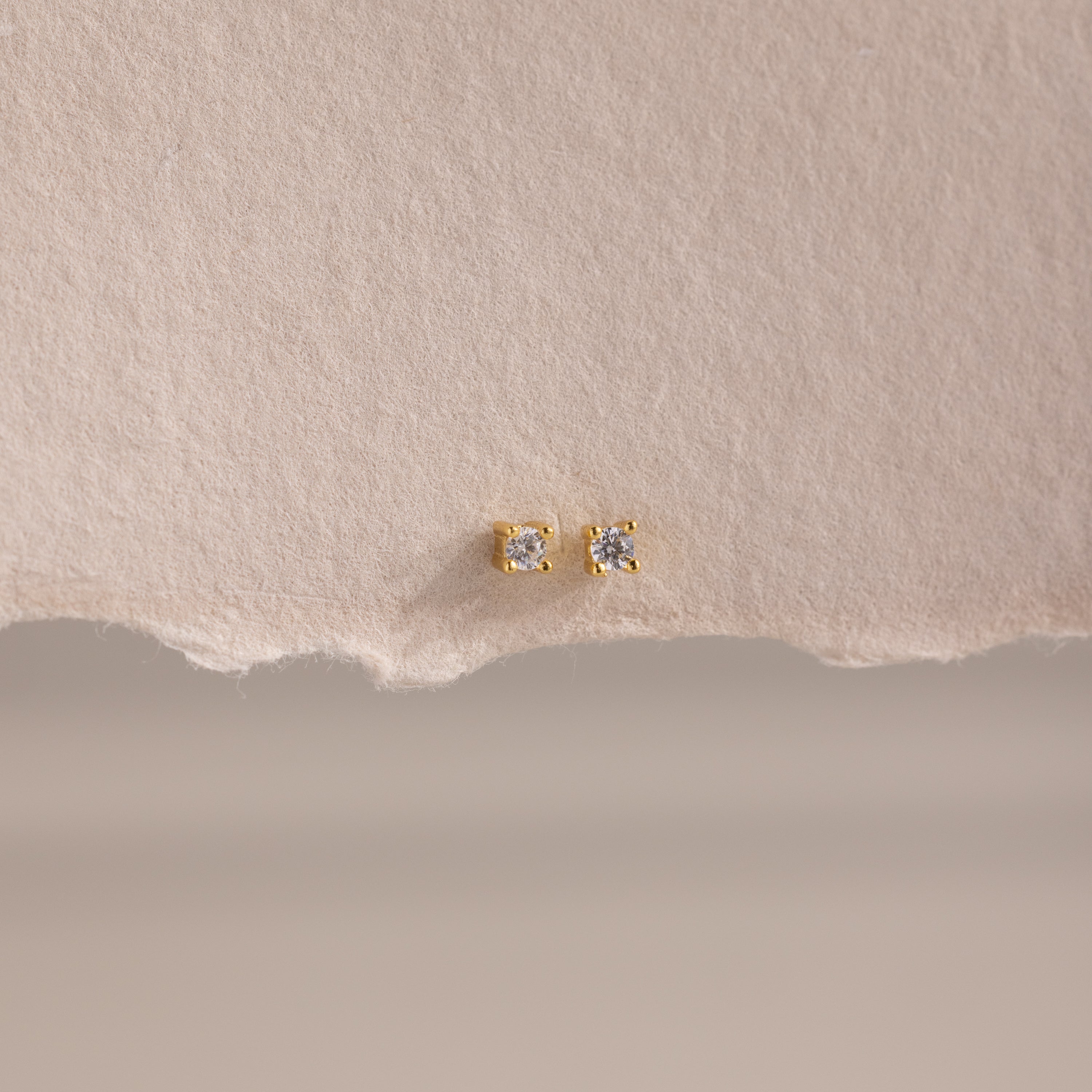 The Dainty Diamond Flatback Studs feature clear gemstones, ideal for multiple piercings, and are shown on a textured beige paper background.
