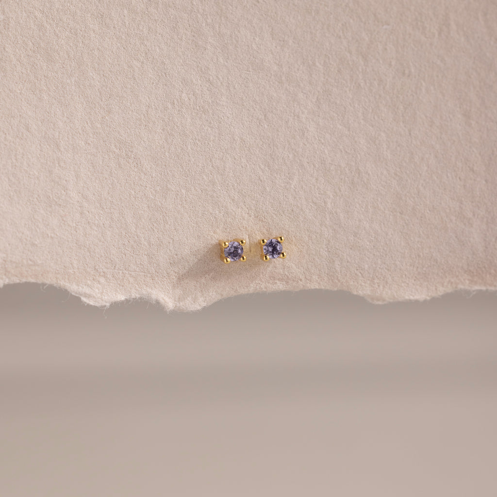 Dainty Alexandrite Flatback Studs—small gold earrings with purple stones, ideal for multiple piercings, shown on a textured, torn piece of beige paper.
