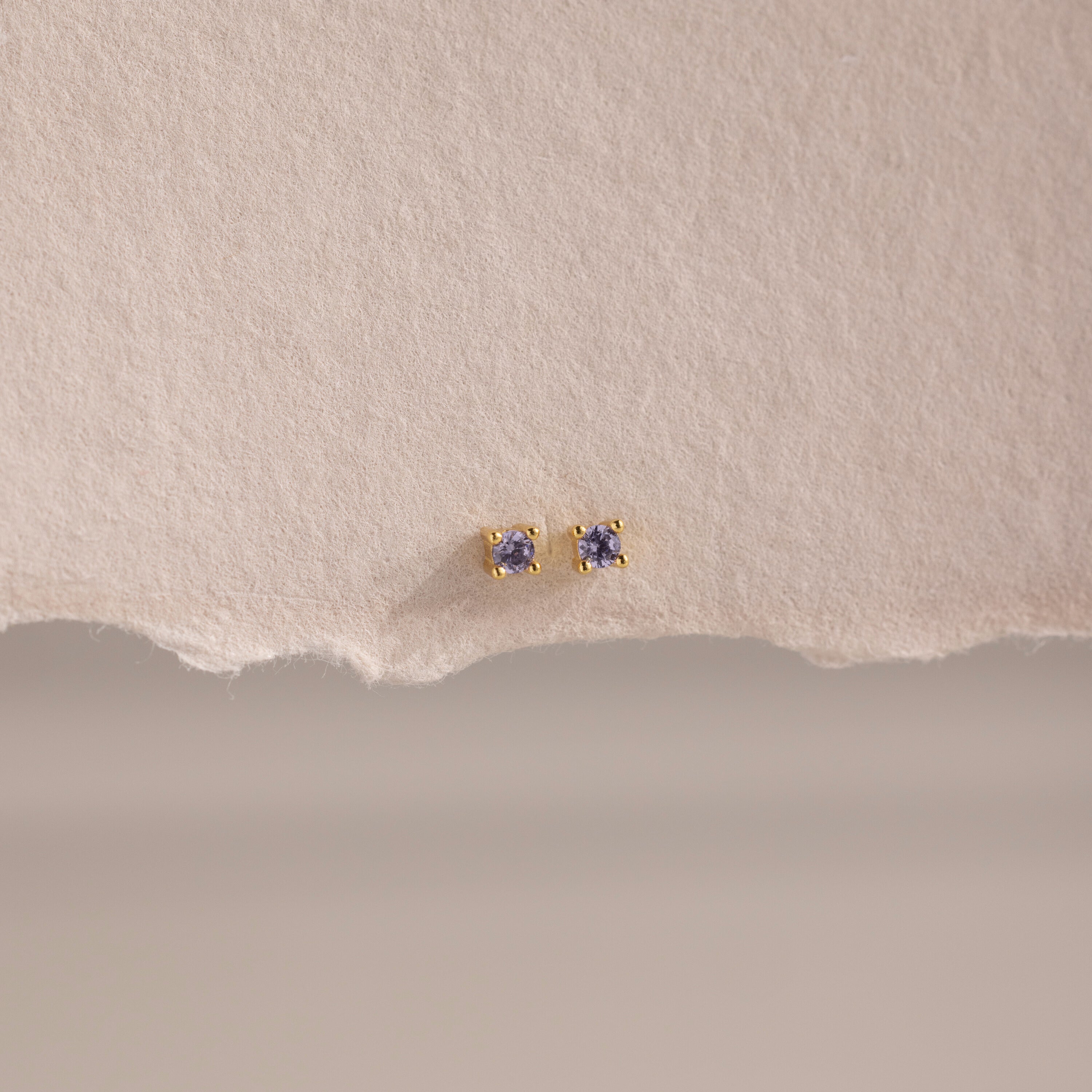 Dainty Alexandrite Flatback Studs—small gold earrings with purple stones, ideal for multiple piercings, shown on a textured, torn piece of beige paper.