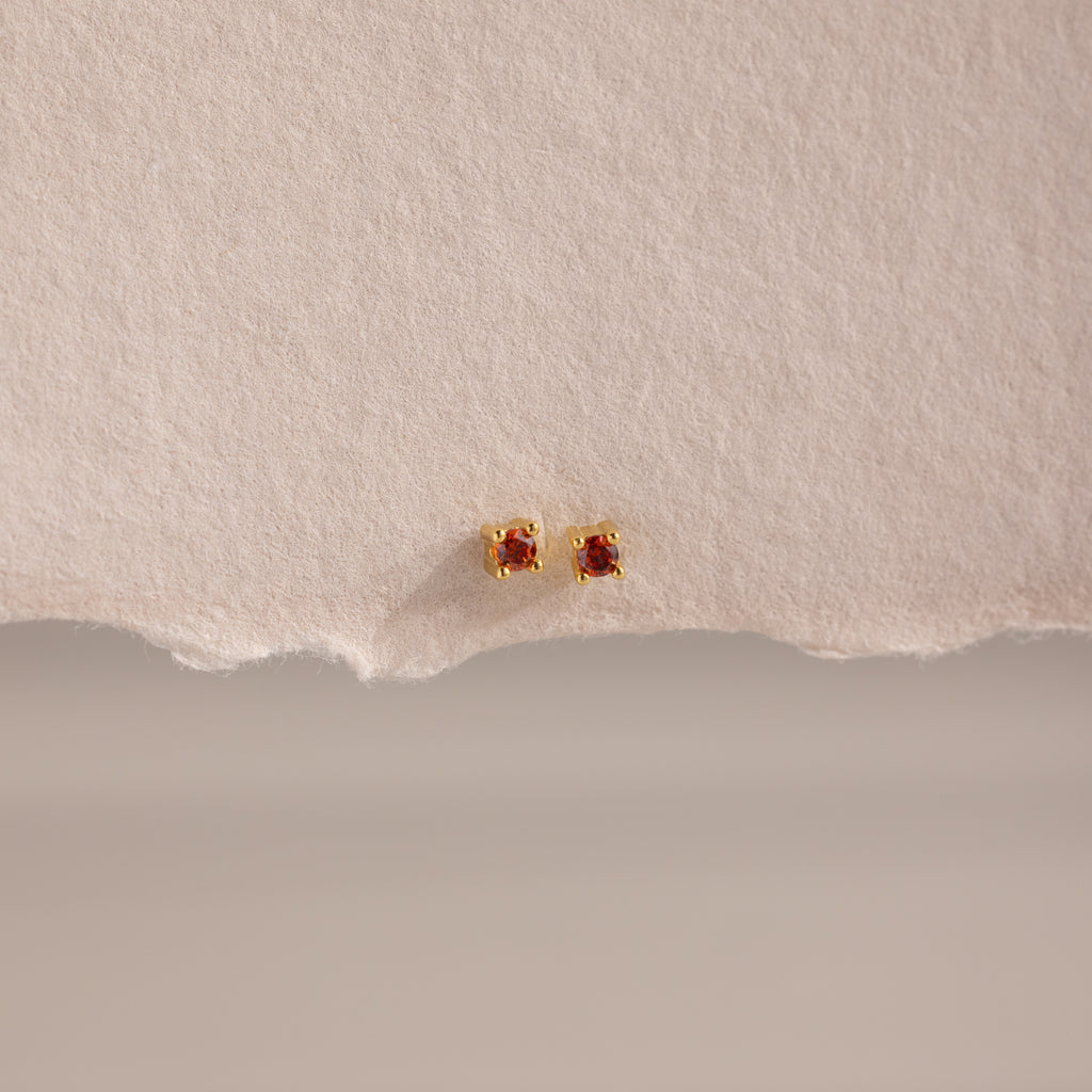 Dainty Garnet Flatback Studs: Gold earrings with small square garnet stones, ideal for multiple piercings. Shown on a textured beige background.