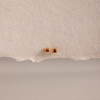 Dainty Garnet Flatback Studs: Gold earrings with small square garnet stones, ideal for multiple piercings. Shown on a textured beige background.