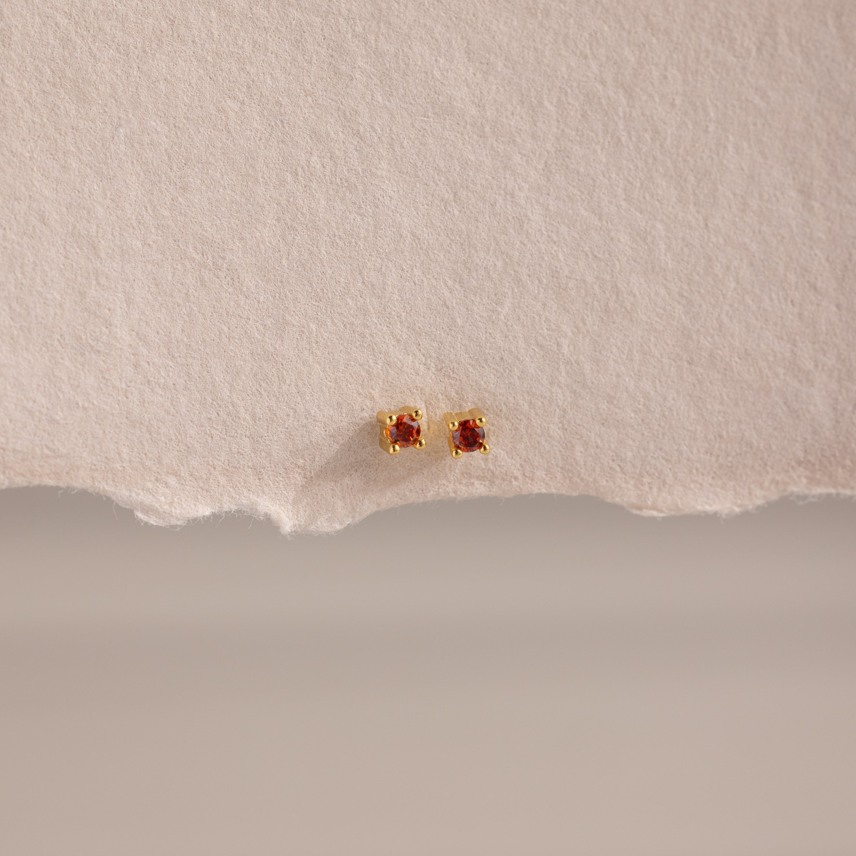 Dainty Garnet Flatback Studs: Gold earrings with small square garnet stones, ideal for multiple piercings. Shown on a textured beige background.