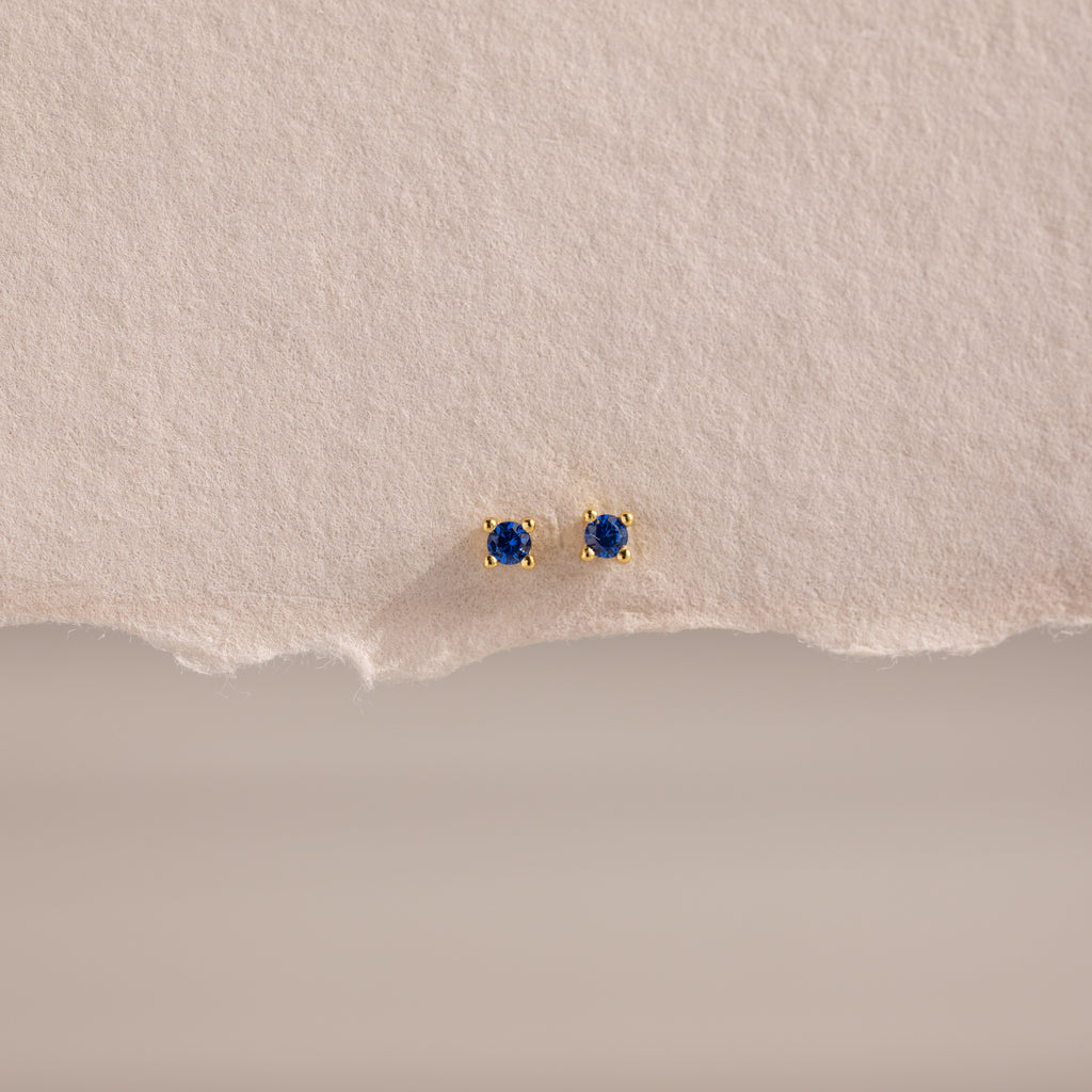 The Dainty Sapphire Flatback Studs feature small round blue gemstones, perfect for multiple piercings, and are displayed on textured beige paper.