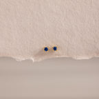 The Dainty Sapphire Flatback Studs feature small round blue gemstones, perfect for multiple piercings, and are displayed on textured beige paper.