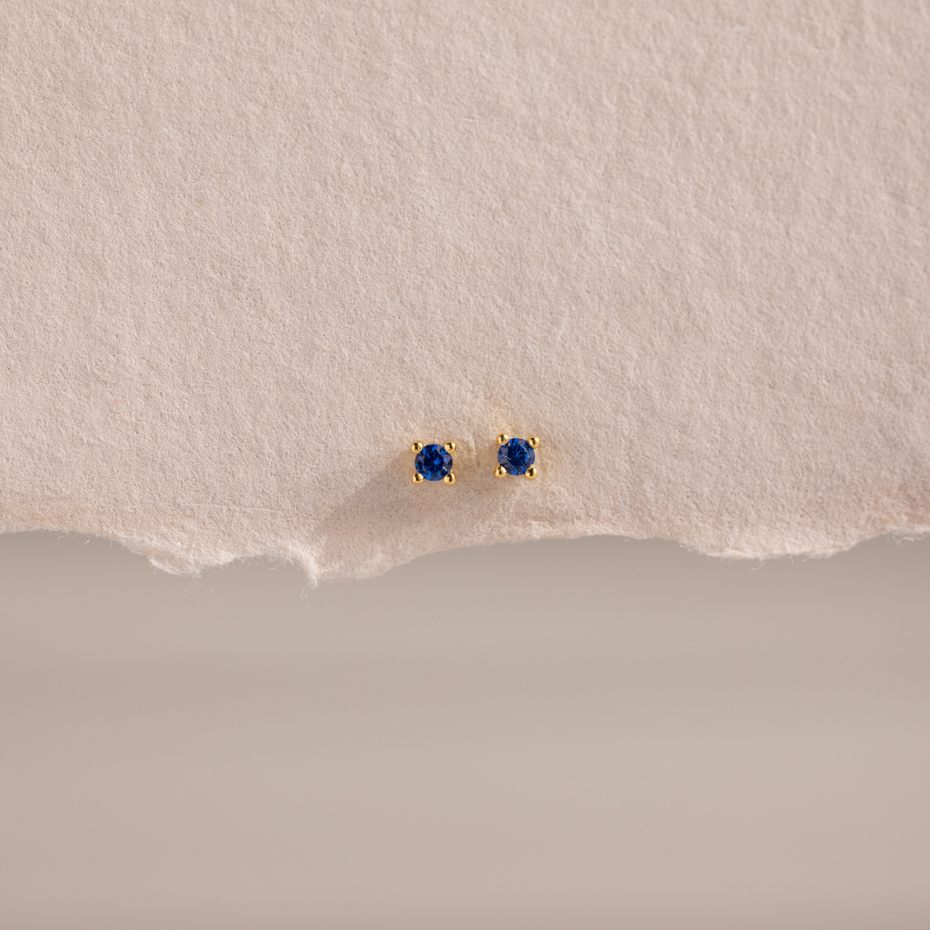 The Dainty Sapphire Flatback Studs feature small round blue gemstones, perfect for multiple piercings, and are displayed on textured beige paper.