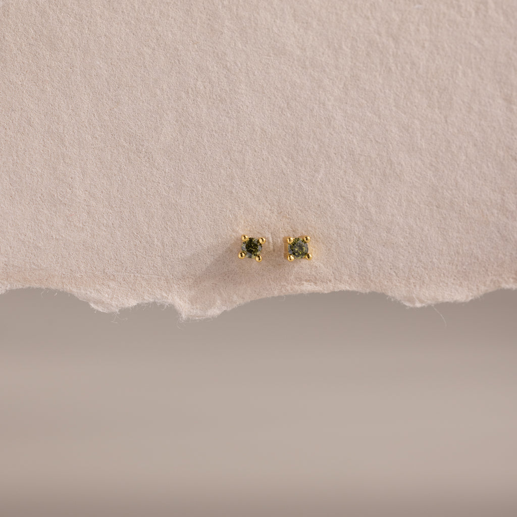 The Dainty Peridot Flatback Studs feature green stones set in gold, displayed on an off-white surface—perfect earrings for multiple piercings.