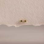 The Dainty Peridot Flatback Studs feature green stones set in gold, displayed on an off-white surface—perfect earrings for multiple piercings.