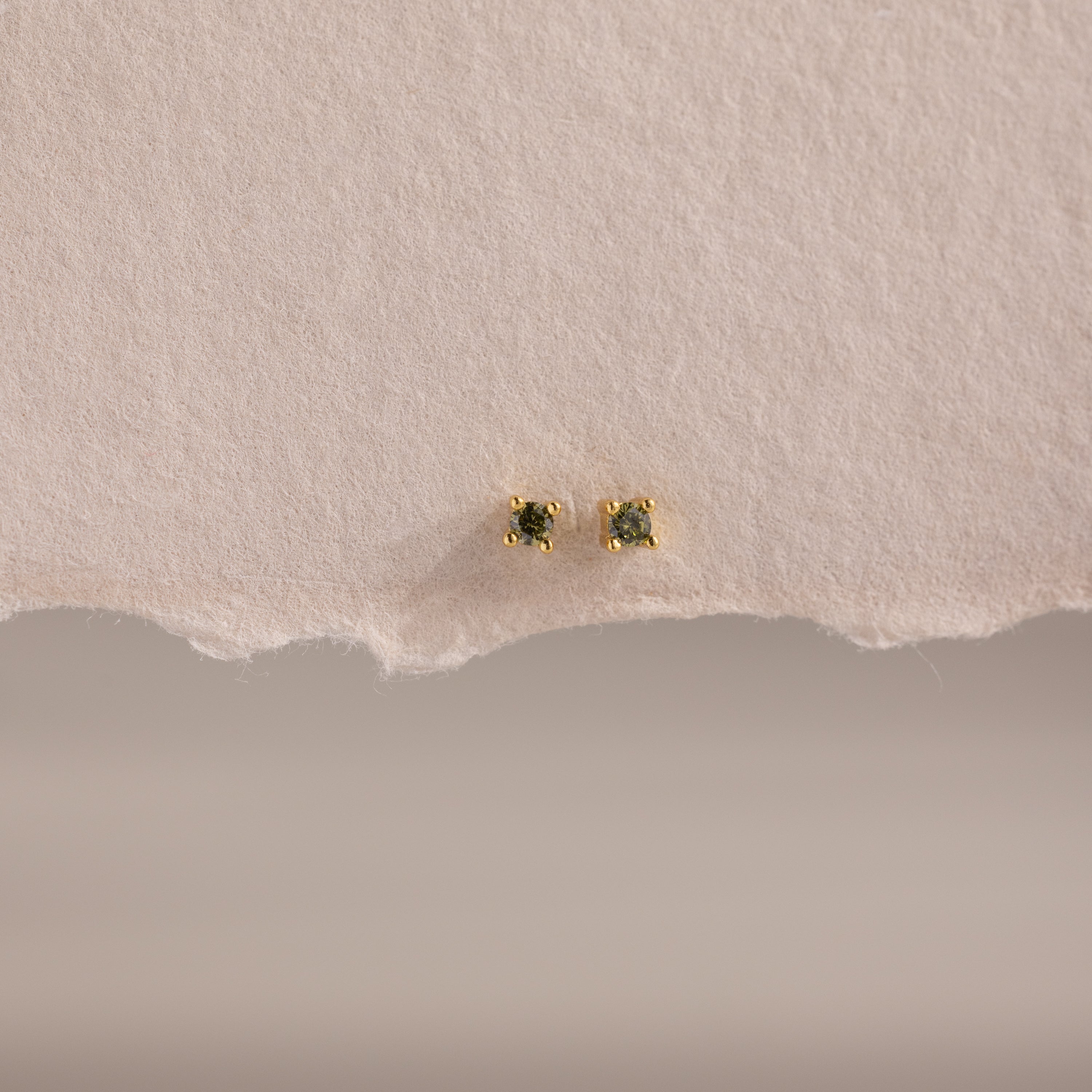 The Dainty Peridot Flatback Studs feature green stones set in gold, displayed on an off-white surface—perfect earrings for multiple piercings.