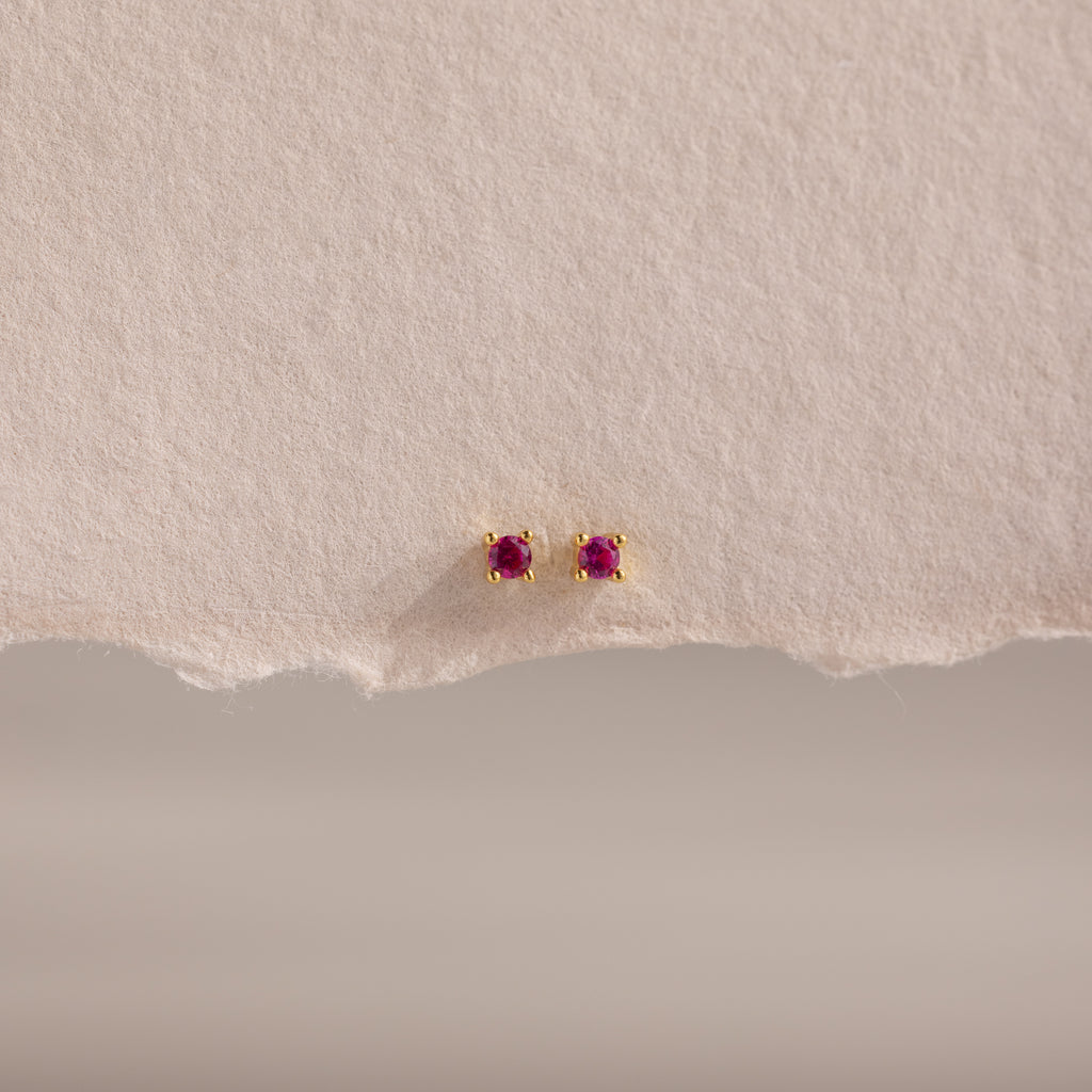 Dainty Ruby Flatback Studs—small gold earrings with round red gemstones, perfect for multiple piercings. Lightweight and ideal for everyday wear, they're stylishly displayed on textured, torn light paper.