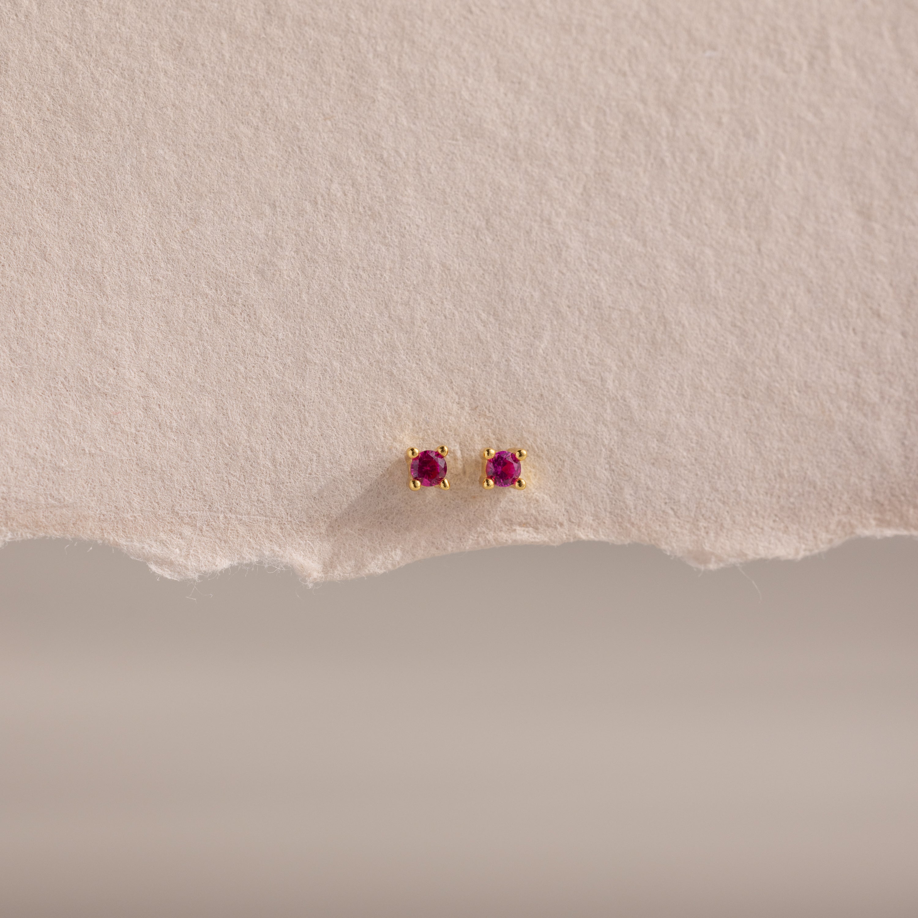 Dainty Ruby Flatback Studs—small gold earrings with round red gemstones, perfect for multiple piercings. Lightweight and ideal for everyday wear, they're stylishly displayed on textured, torn light paper.