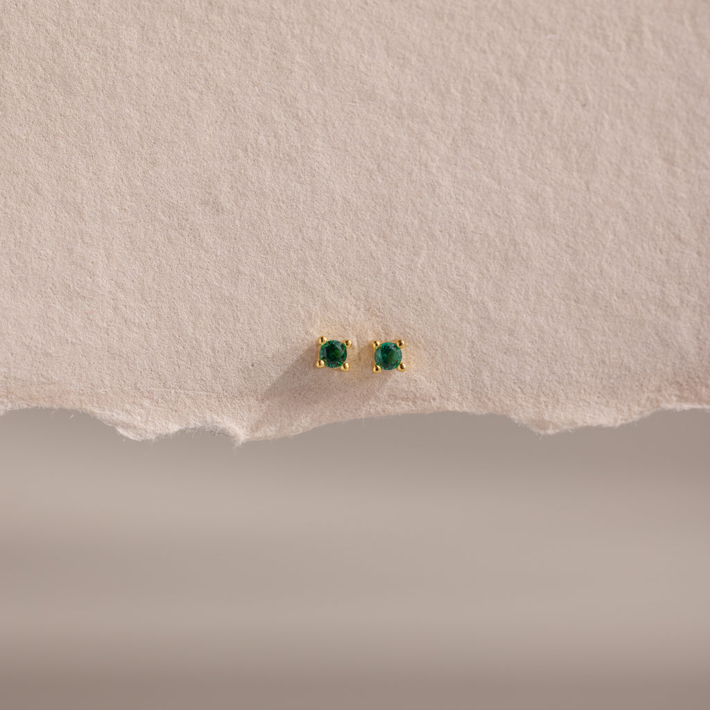 Dainty Emerald Flatback Studs are small gold earrings with green gemstones, displayed on textured beige paper. Lightweight, they're ideal for multiple piercings and add a subtle pop of color to any look.