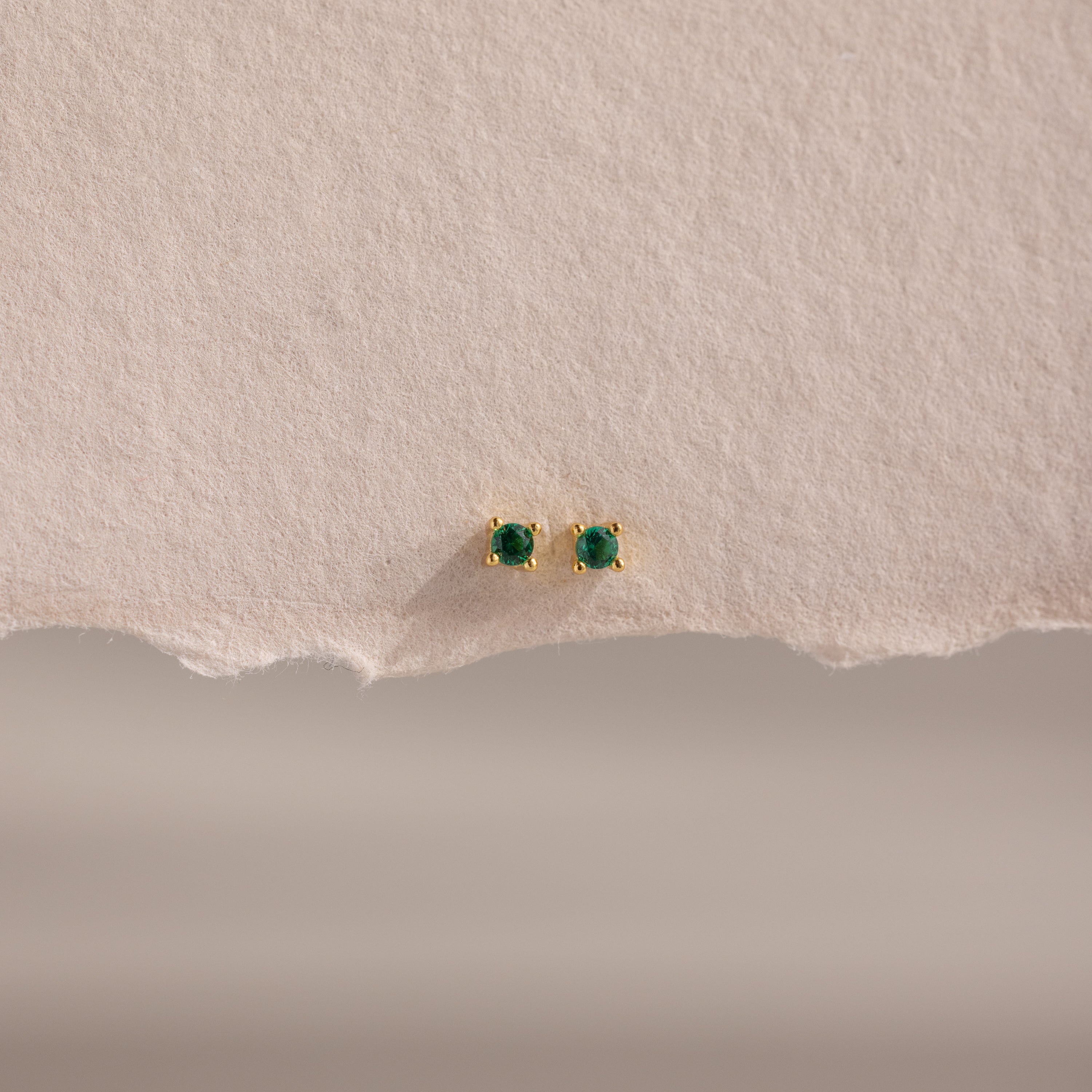Dainty Emerald Flatback Studs are small gold earrings with green gemstones, displayed on textured beige paper. Lightweight, they're ideal for multiple piercings and add a subtle pop of color to any look.