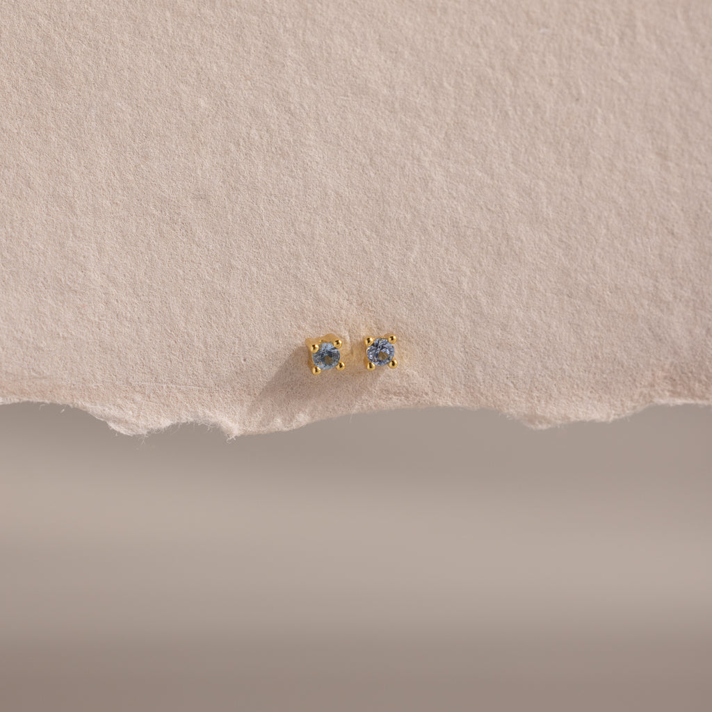 Dainty Aquamarine Flatback Studs with small round aquamarines, perfect for multiple piercings, displayed on textured, torn-edge beige paper.