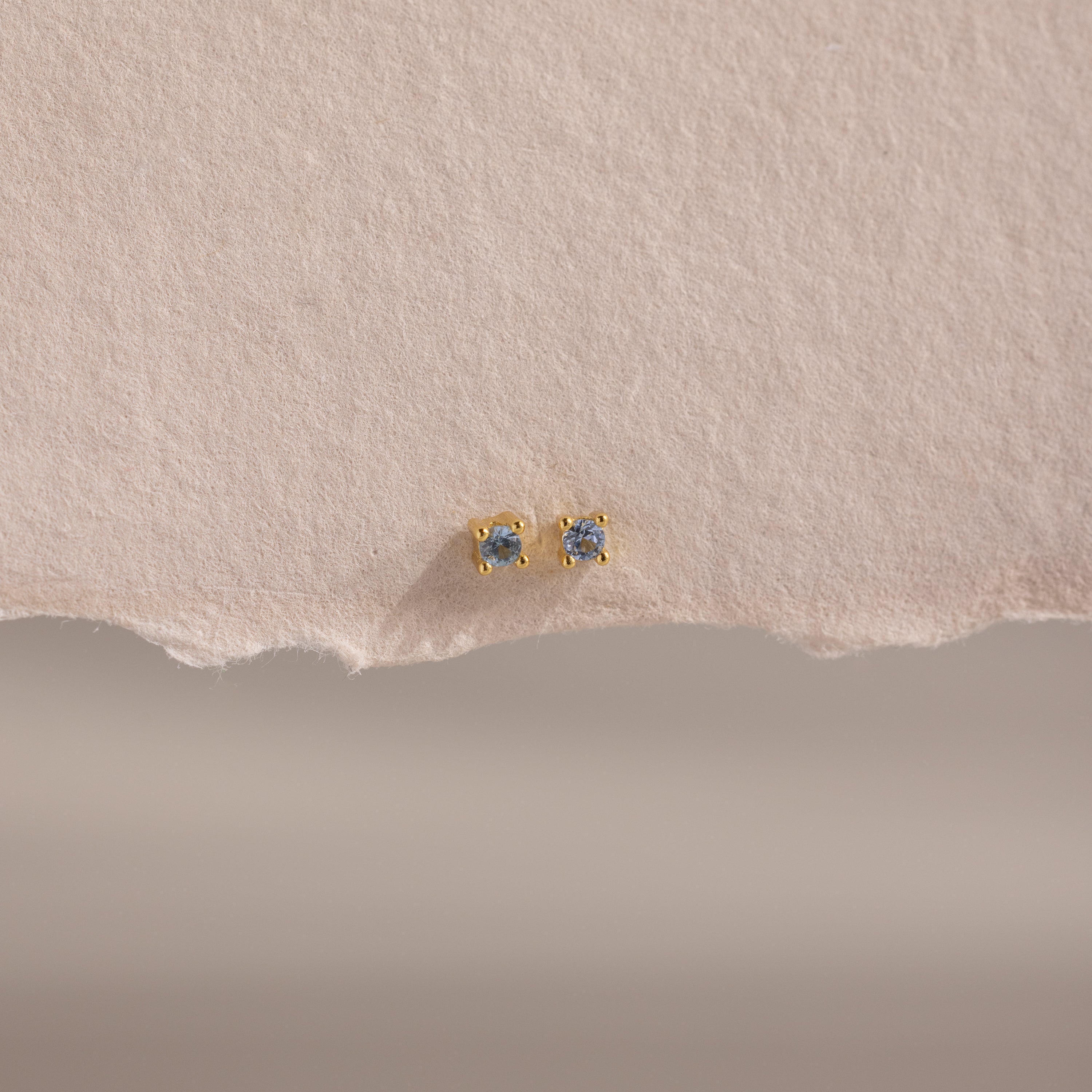 Dainty Aquamarine Flatback Studs with small round aquamarines, perfect for multiple piercings, displayed on textured, torn-edge beige paper.