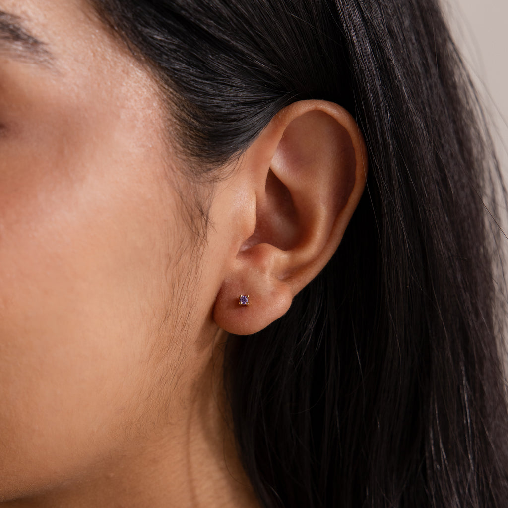 Close-up of a woman's ear with straight dark hair, wearing Dainty Birthstone Flatback Studs featuring a purple gemstone—a timeless look ideal for multiple piercings.