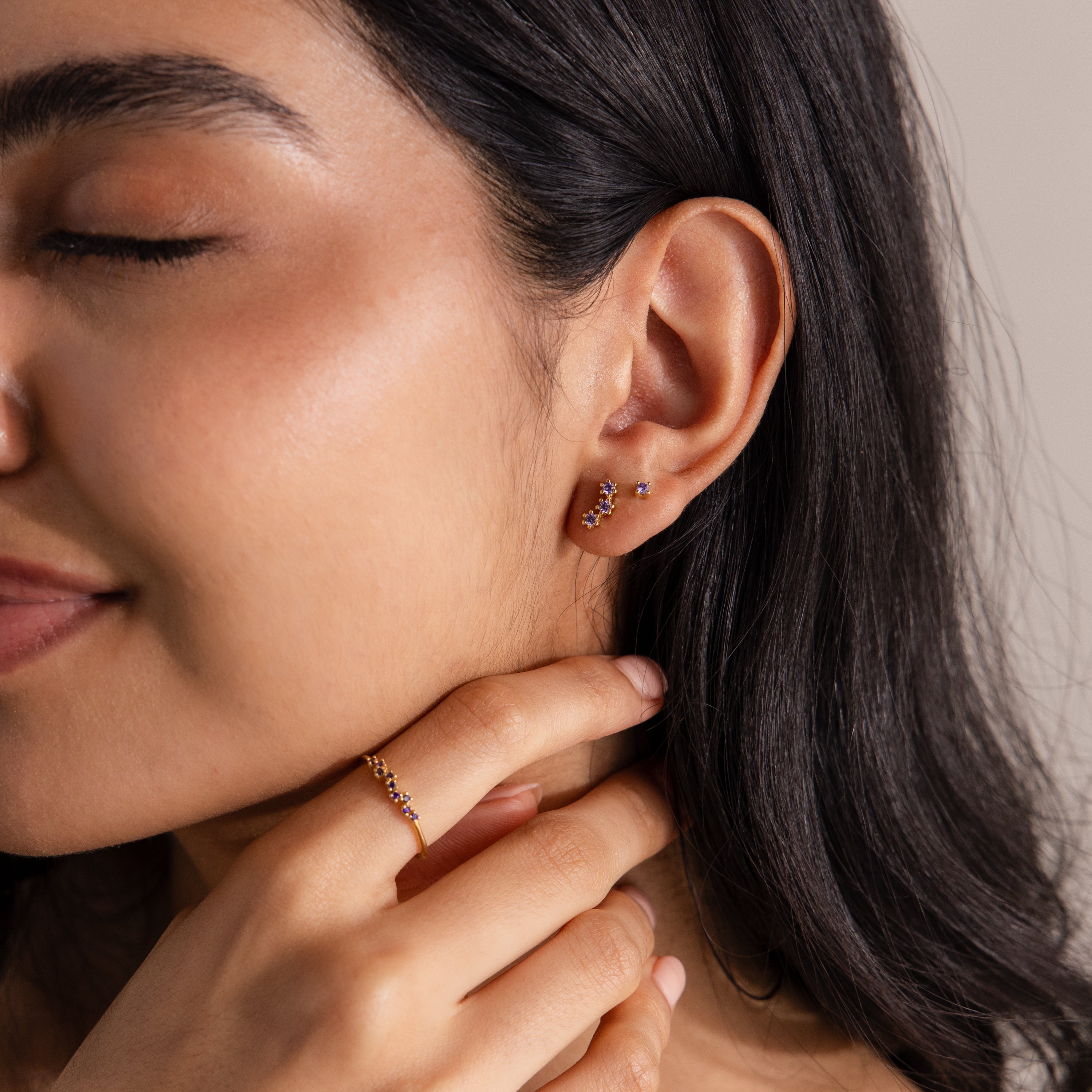 With closed eyes, a woman touches her neck, wearing timeless gold earrings, a matching ring, and multiple Dainty Birthstone Flatback Studs adorning her piercings.