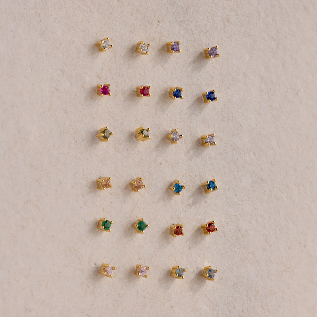 Twenty pairs of Dainty Amethyst Flatback Studs, ideal for multiple piercings, are displayed in a neat grid on a beige surface.