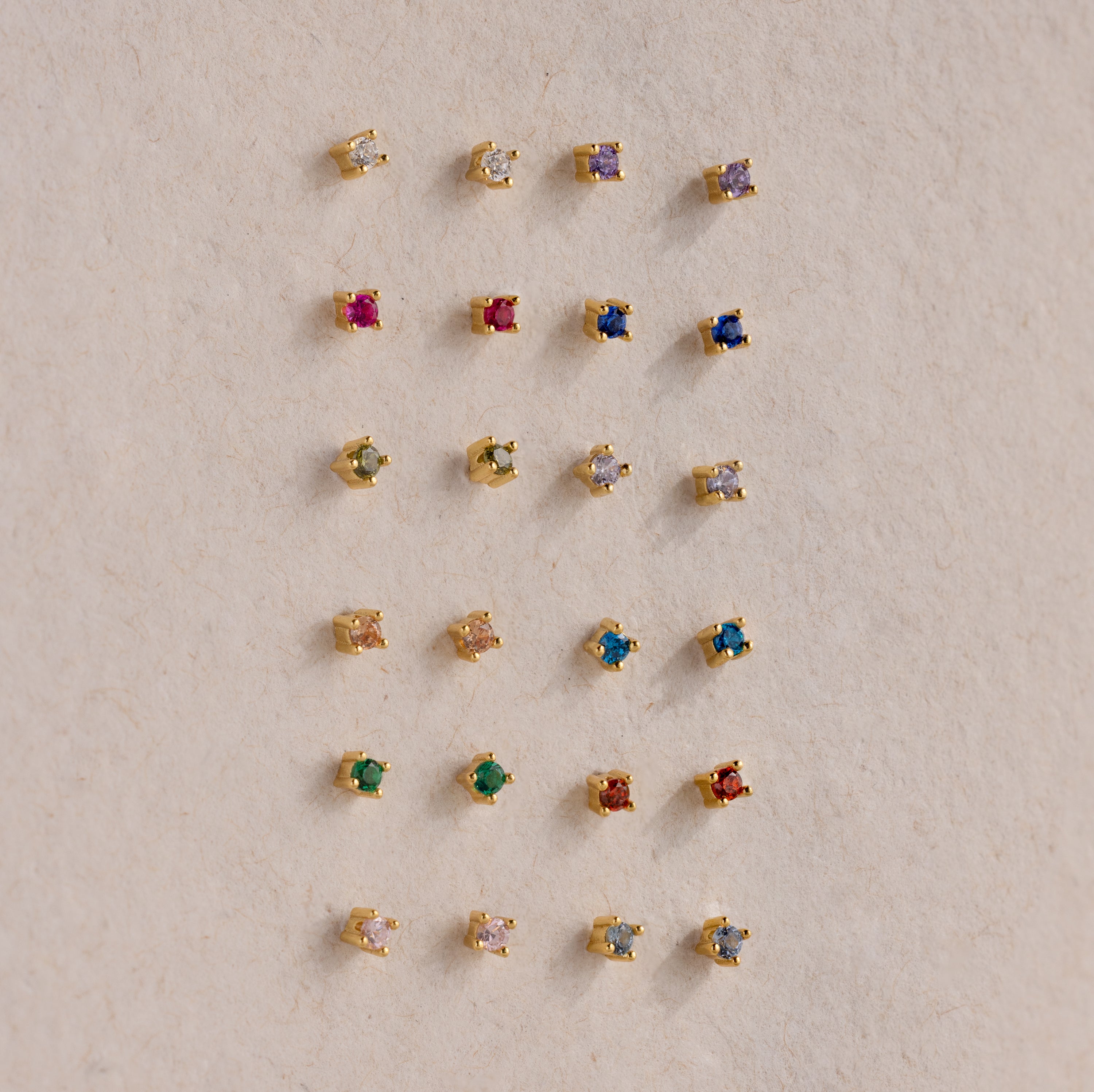 Nine pairs of Dainty Birthstone Flatback Studs, each with a unique colored stone, are displayed in a grid on a beige surface, creating a timeless style ideal for multiple piercings.