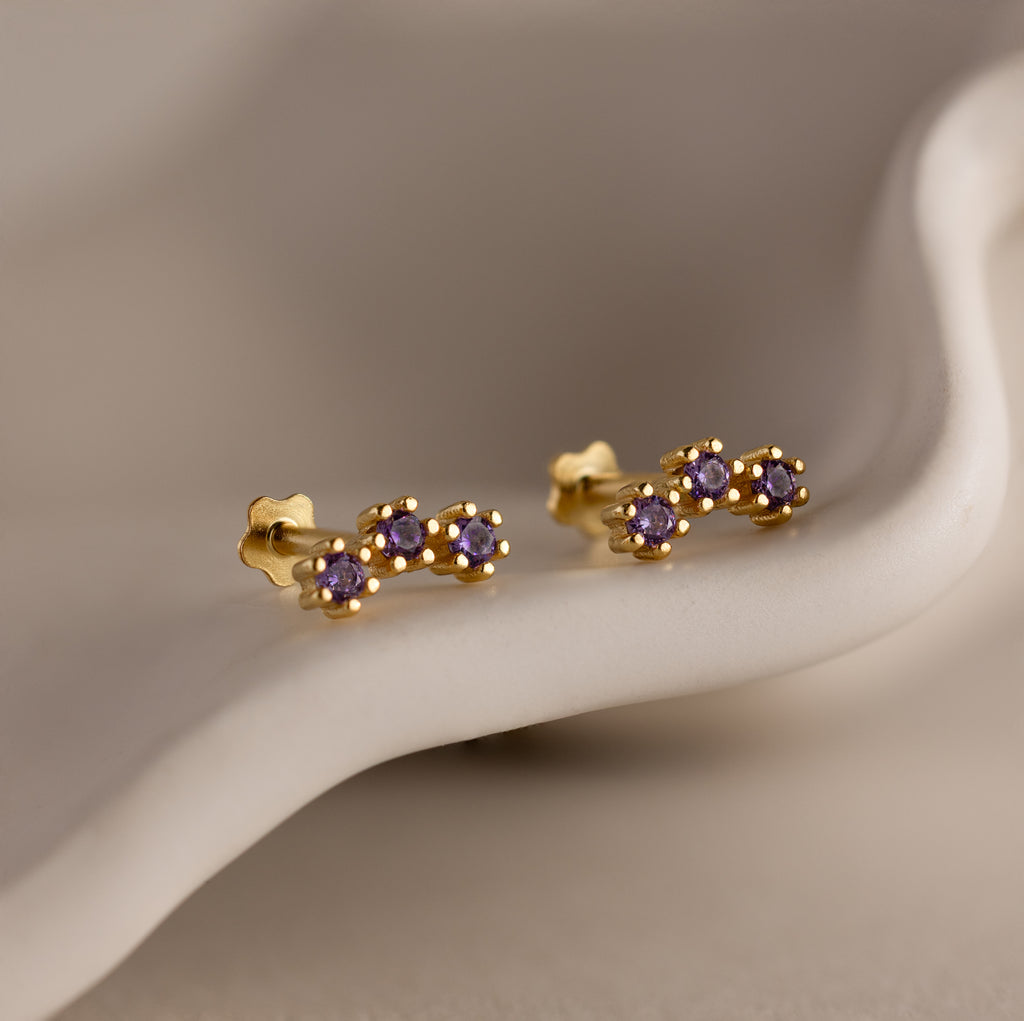 Birthstone Cluster Flatback Studs—gold earrings featuring three small purple gemstones—are perfect for an elegant stack and are displayed on a smooth, neutral surface.