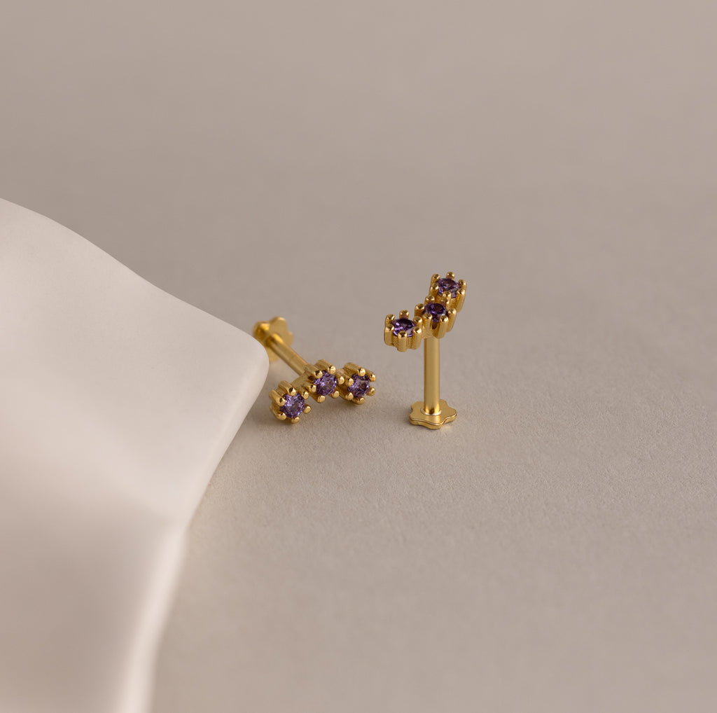 Two Birthstone Cluster Flatback Studs with small purple gemstones rest on a light beige surface beside white fabric—ideal accents to elevate your earring stack.