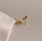 Two Birthstone Cluster Flatback Studs with small purple gemstones rest on a light beige surface beside white fabric—ideal accents to elevate your earring stack.