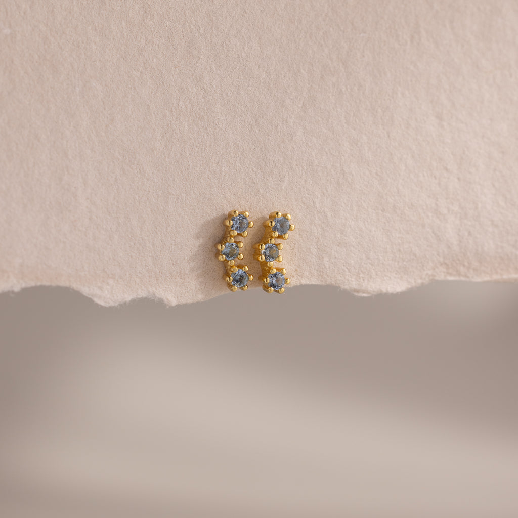 Aquamarine Cluster Flatback Studs: small gold earrings with blue gemstones, shown on a textured light beige surface. Elegant birthstone studs, perfect for any occasion.