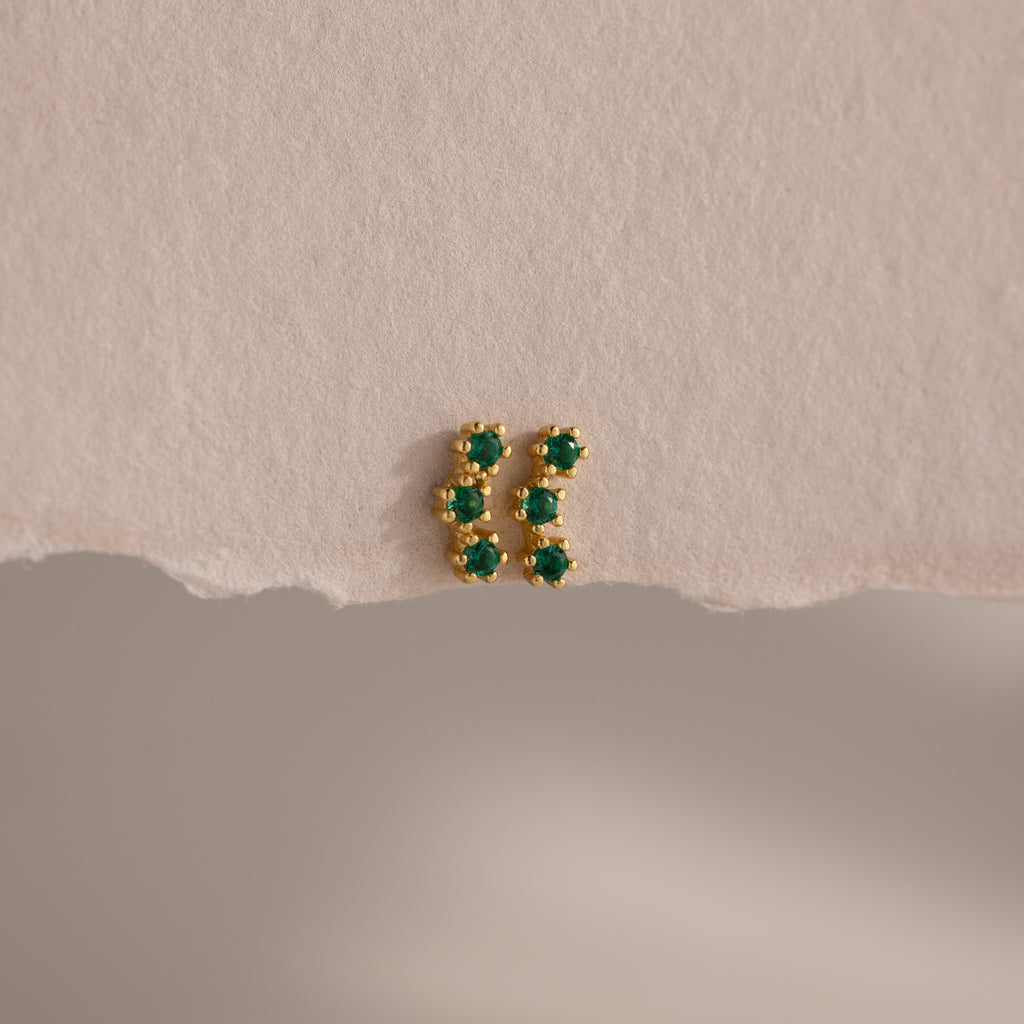 Emerald Cluster Flatback Studs: small gold earrings featuring six green stones in two vertical rows, shown on a textured beige surface.