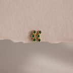 Emerald Cluster Flatback Studs: small gold earrings featuring six green stones in two vertical rows, shown on a textured beige surface.