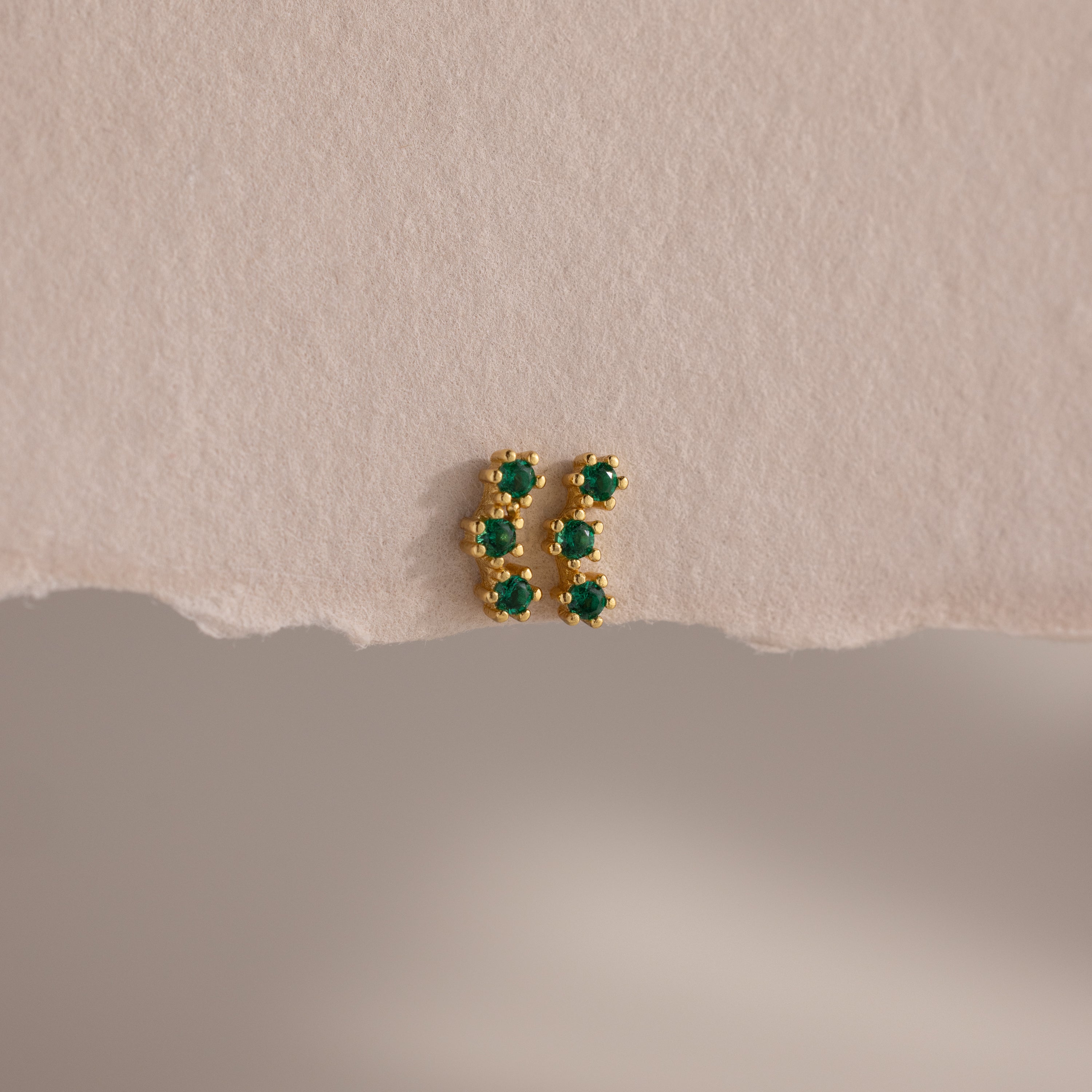 Emerald Cluster Flatback Studs: small gold earrings featuring six green stones in two vertical rows, shown on a textured beige surface.
