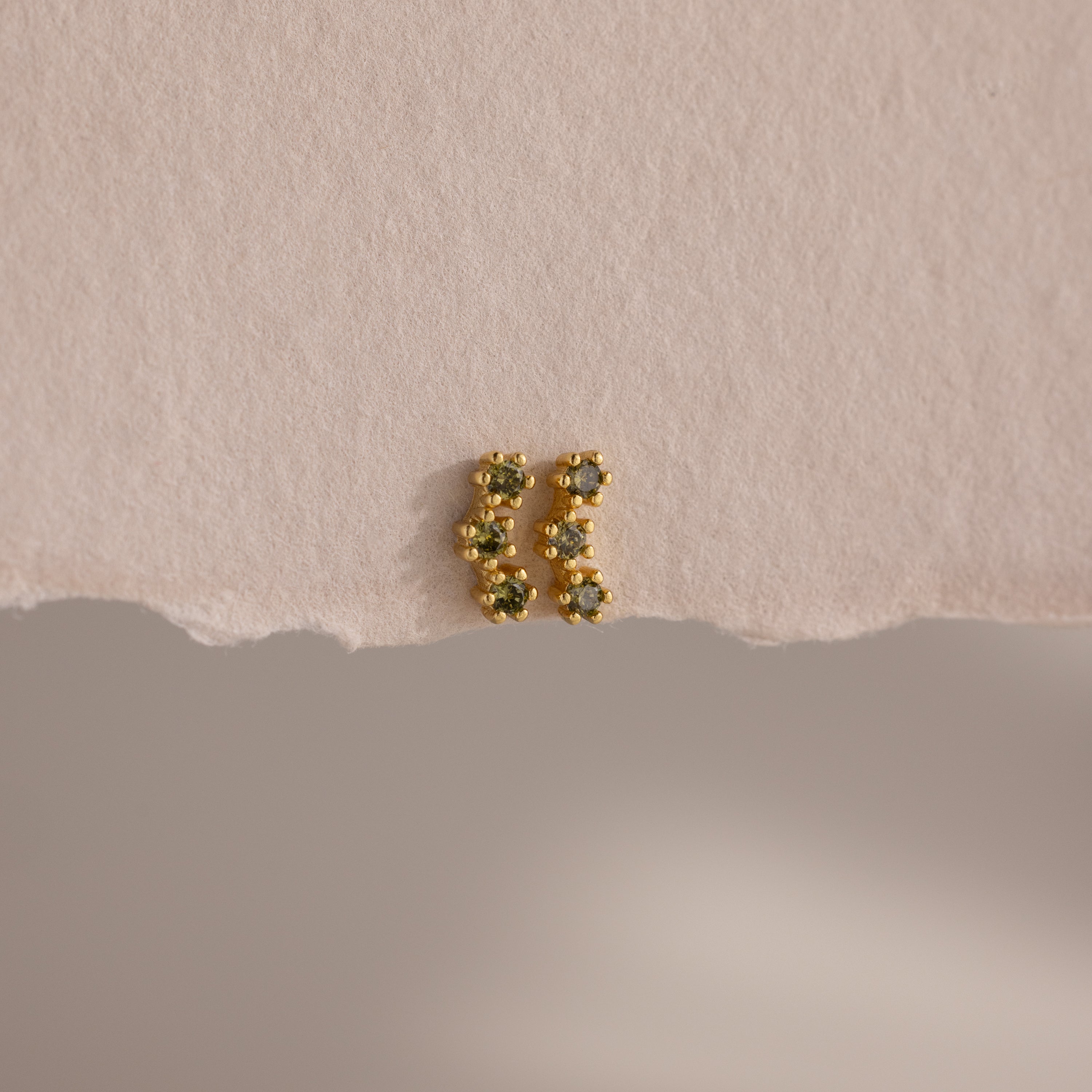 A pair of Peridot Cluster Flatback Studs with green gemstones rests elegantly on a textured, off-white surface.